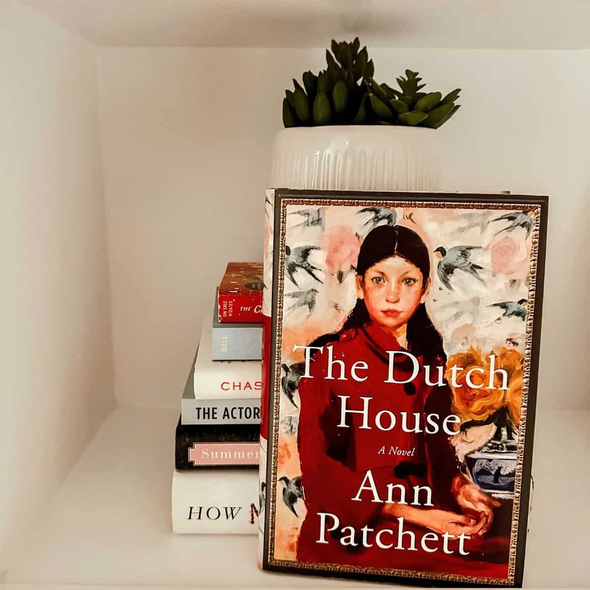 the dutch house by ann patchett on a bookshelf with books and a plant.