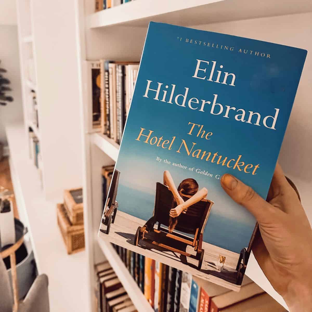 the hotel nantucket by elin hilderbrand held up in front of a bookcase.