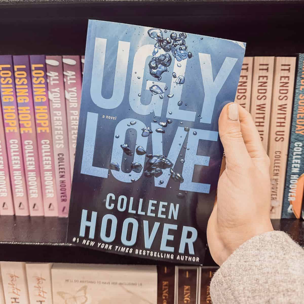 Ugly Love by Colleen Hoover held in front of a bookshelf with other Colleen Hoover books.