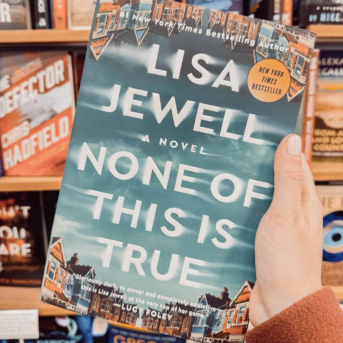 None of this is true by Lisa Jewell held in front of a bookshelf.